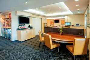 A hotel lobby with a reception desk, a dining area with a round table and chairs, a TV mounted on the wall, and a patterned carpet. - TownePlace Suites by Marriott Baton Rouge Gonzales (Gonzales)