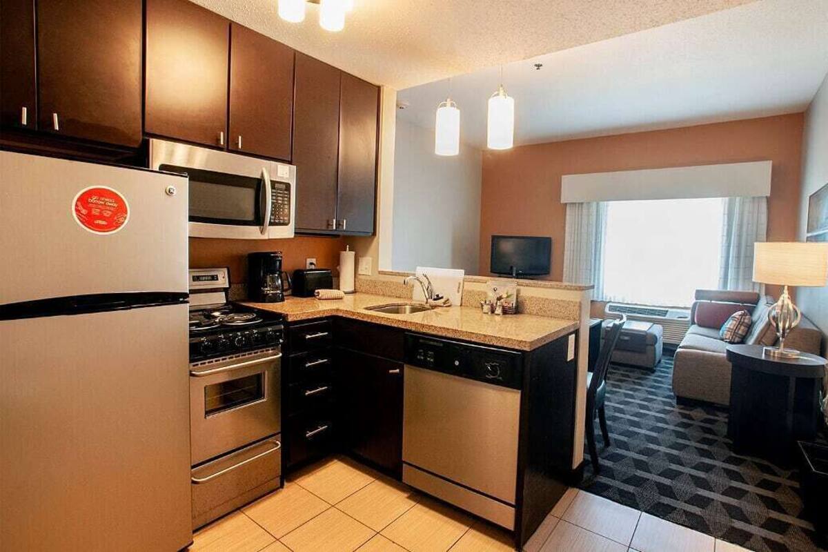 A compact hotel kitchen with stainless steel appliances, a flat-screen TV, and a seating area with a sofa and lamp.