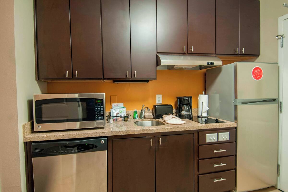 A compact kitchen with dark brown cabinets, a stainless steel microwave, a dishwasher, a coffee maker, and a refrigerator.