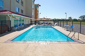 A rectangular pool with a tiled deck, surrounded by blue lounge chairs and red umbrellas. - TownePlace Suites by Marriott Baton Rouge Gonzales (Gonzales)