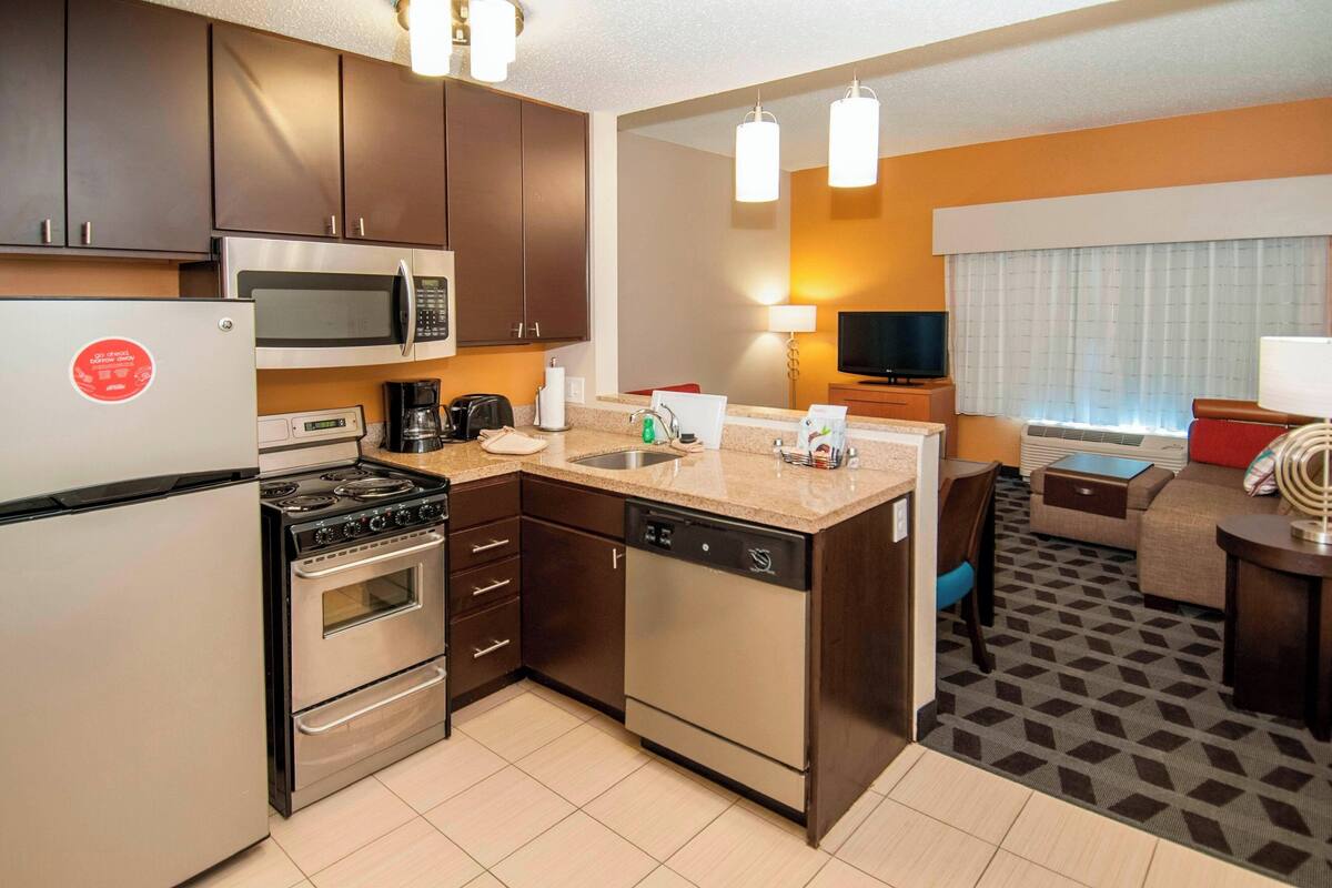 A modern kitchen with stainless steel appliances, a built-in microwave, and a dishwasher. There is a dining area with a table and chairs, and a TV mounted on the wall.