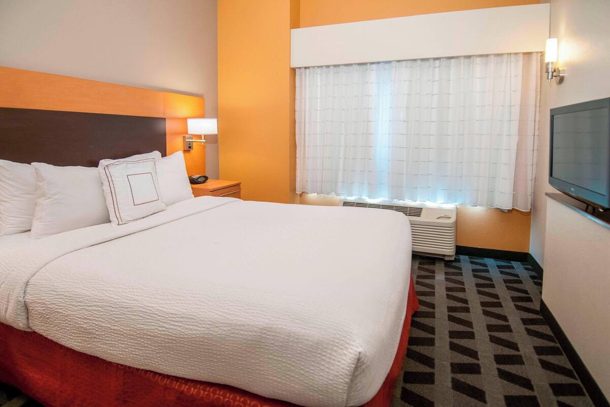 A hotel room with a large bed, a bedside table with a lamp, a window with curtains, and a television on the wall.