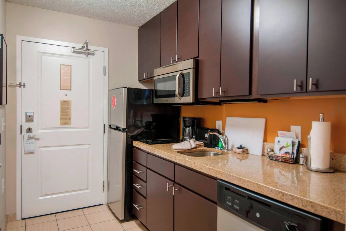 A compact kitchen with dark brown cabinets, a stainless steel microwave, a coffee maker, and a dishwasher.
