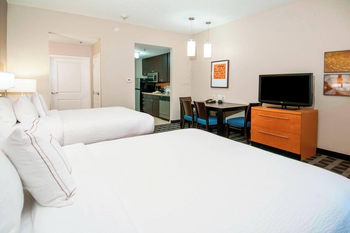 A hotel room with two beds, a desk with a TV, a chair, and a small kitchenette.