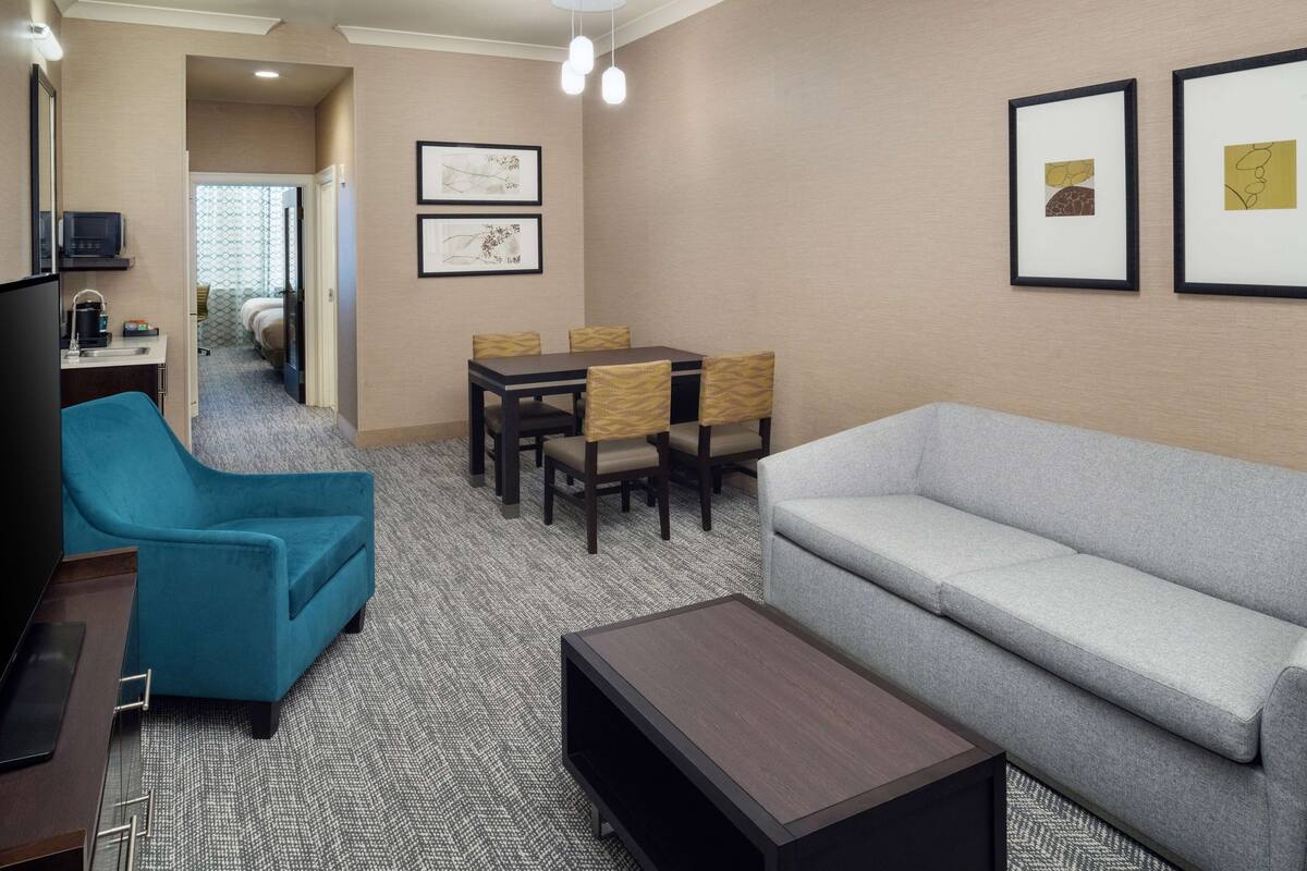 Habitación de hotel con sofá, un sillón azul, una mesa de comedor con sillas, cafetera y cuadros en la pared.
