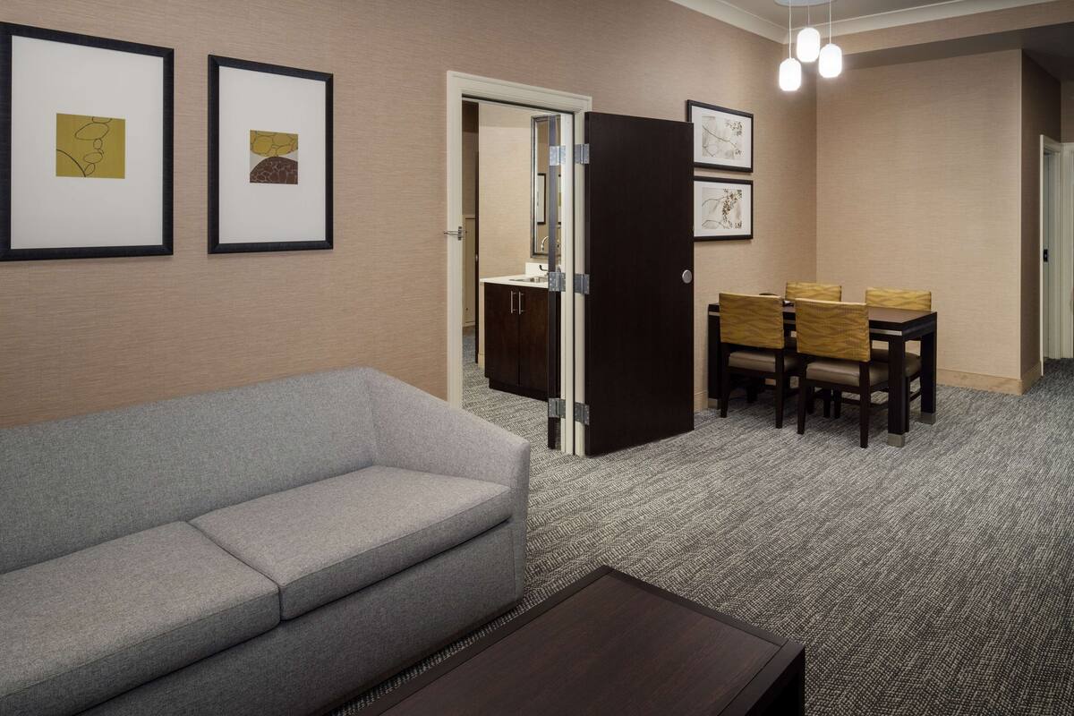 Habitación de hotel con un sofá gris, una mesa de comedor con cuatro sillas y obras de arte enmarcadas en las paredes.