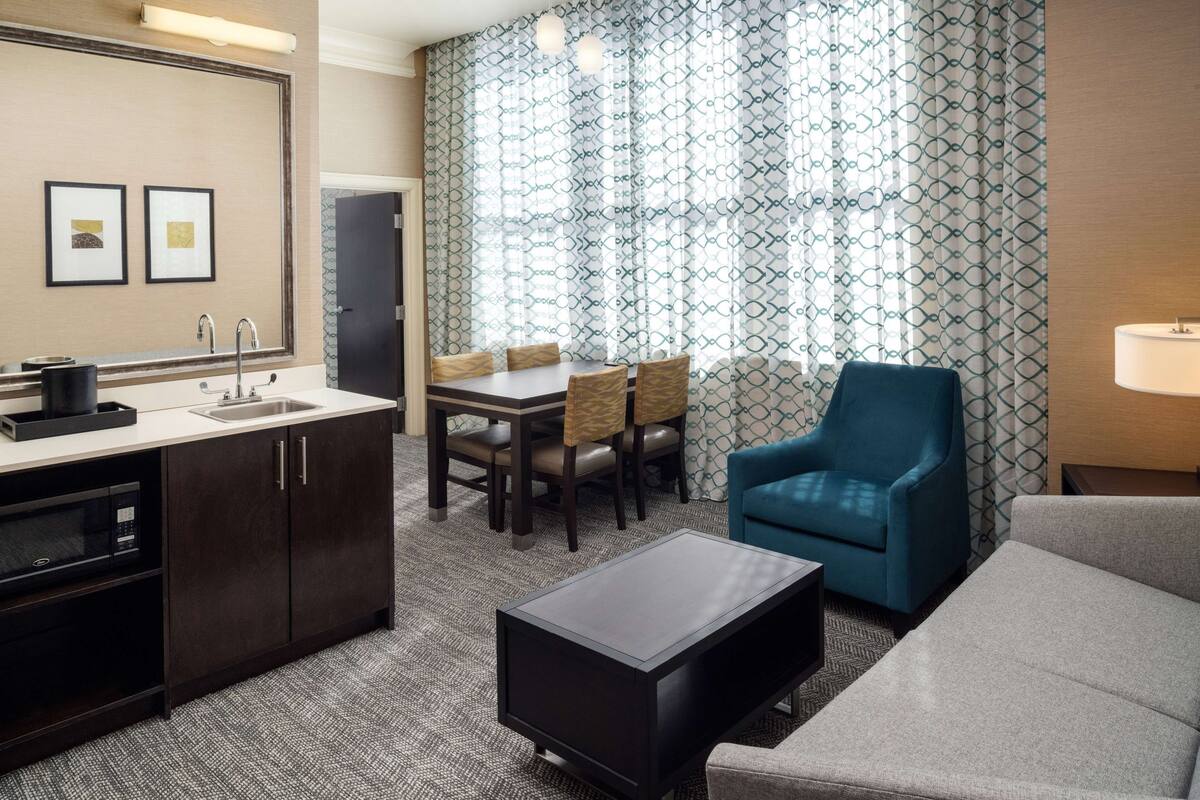 Una habitación de hotel con kitchenette, una zona de comedor con mesa y sillas, un sillón azul, un sofá gris y una lámpara.