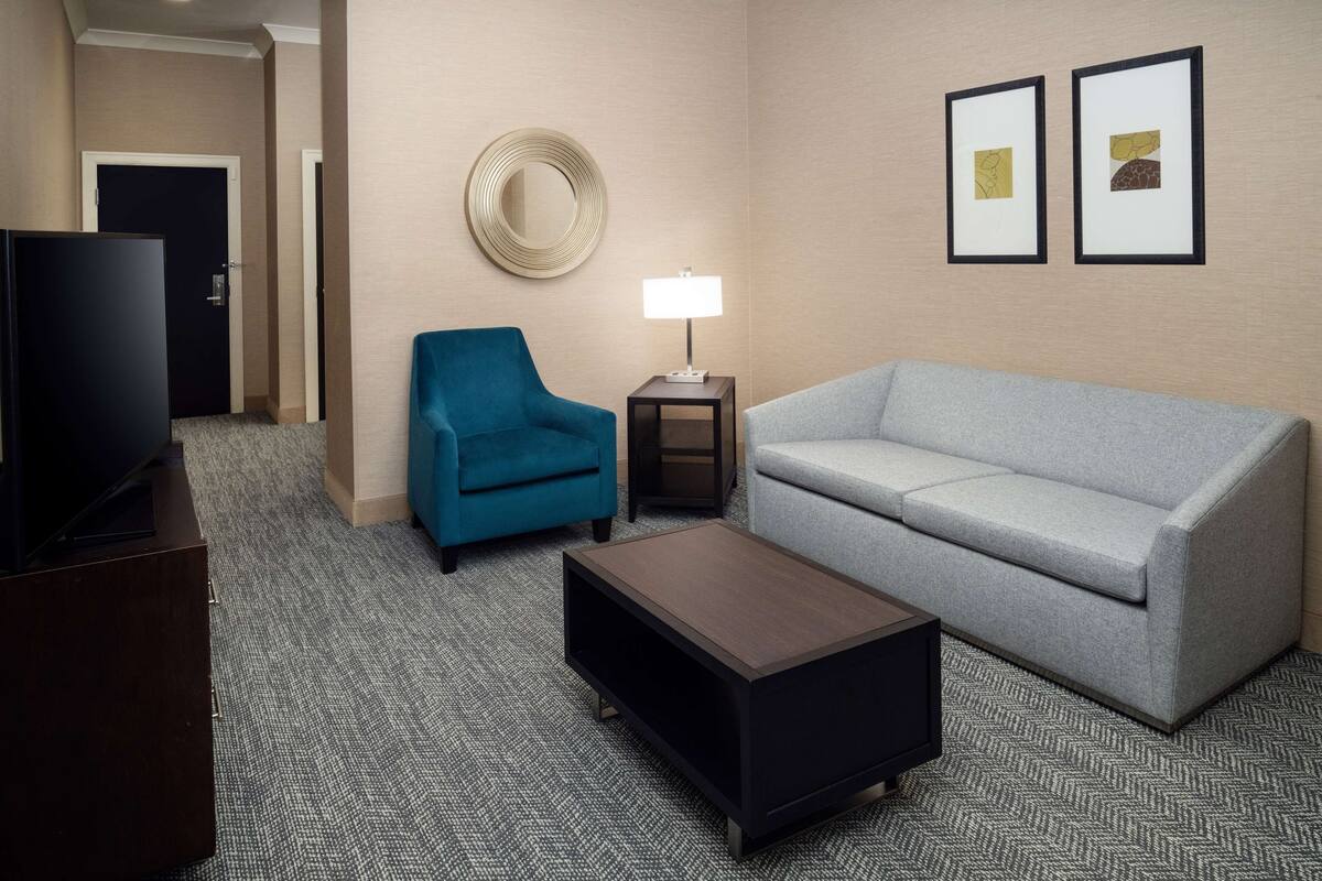 Habitación de hotel con sofá gris, sillón azul, una mesita, televisor, lámpara y dos cuadros en la pared.