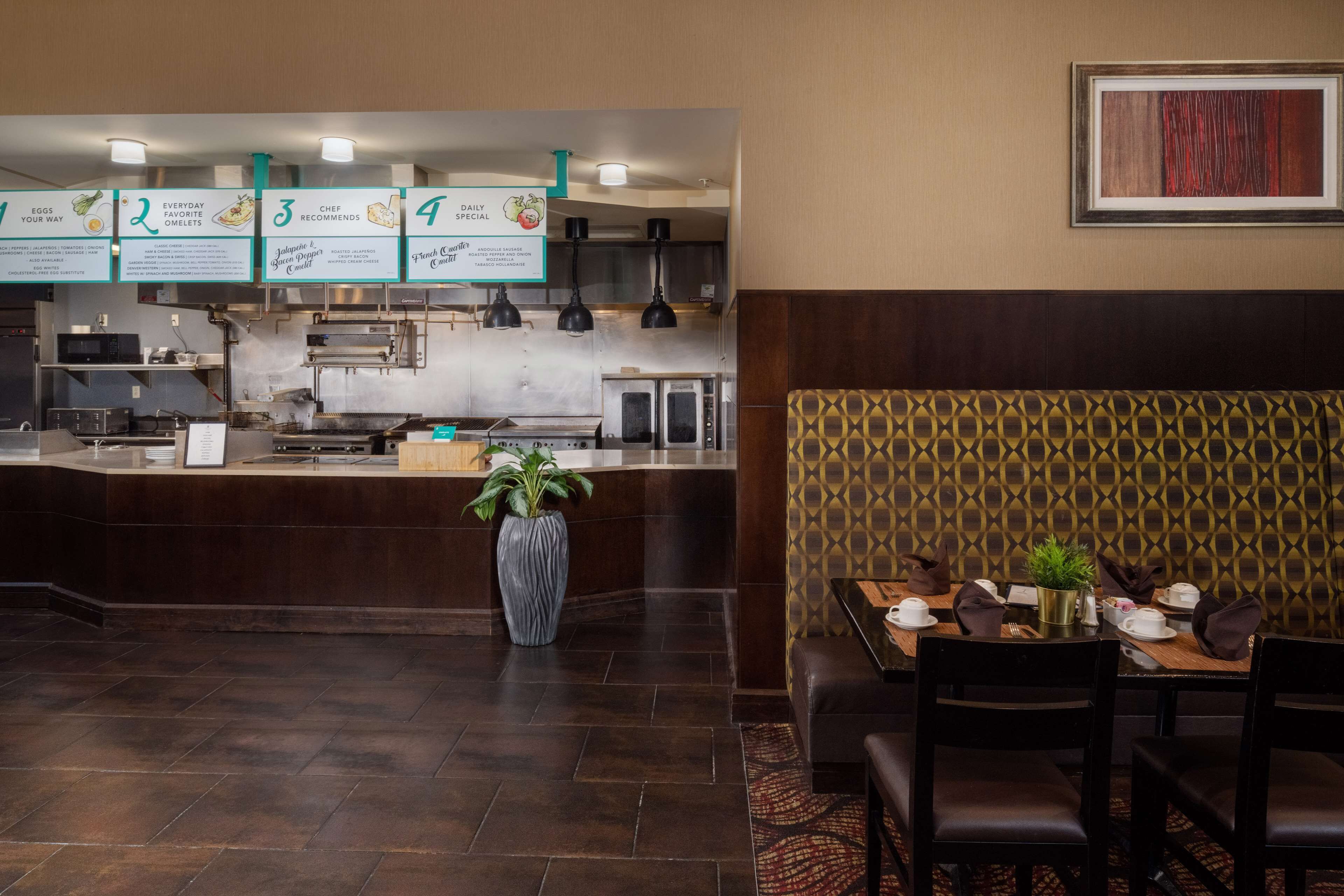 Un restaurant avec un comptoir, des panneaux affichant le menu, un espace où s’asseoir avec un mur décoré de motifs, et une plante en pot.