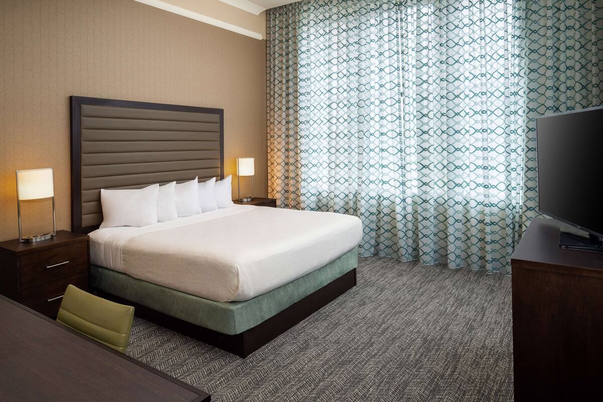 Una habitación de hotel con una cama grande, dos mesitas de noche con lámparas, un televisor y cortinas estampadas.