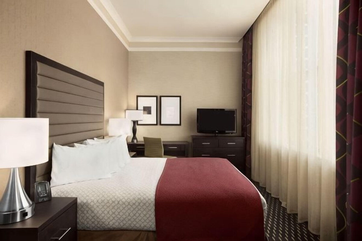 Quarto de hotel com cama, televisão, mesa de cabeceira e escrivaninha.