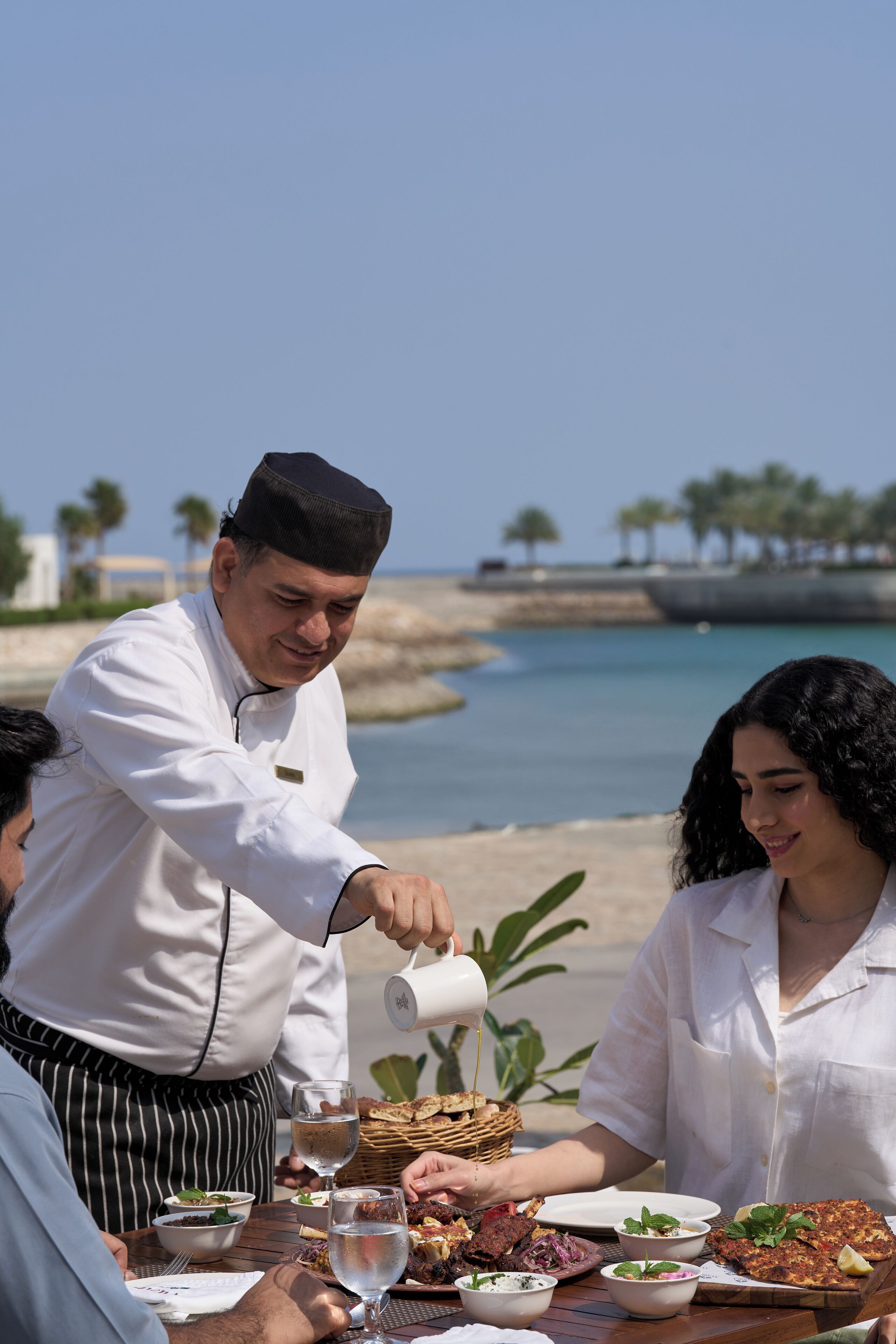 A chef serving food at an outdoor dining table with a view of the sea and palm trees.