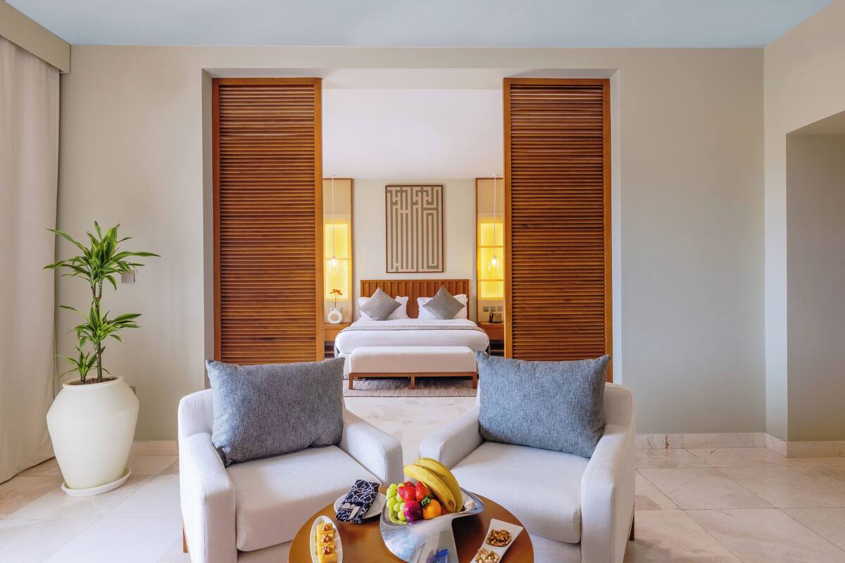 Une chambre d’hôtel moderne avec un grand lit, deux canapés blancs, une table basse avec des fruits et une plante en pot.