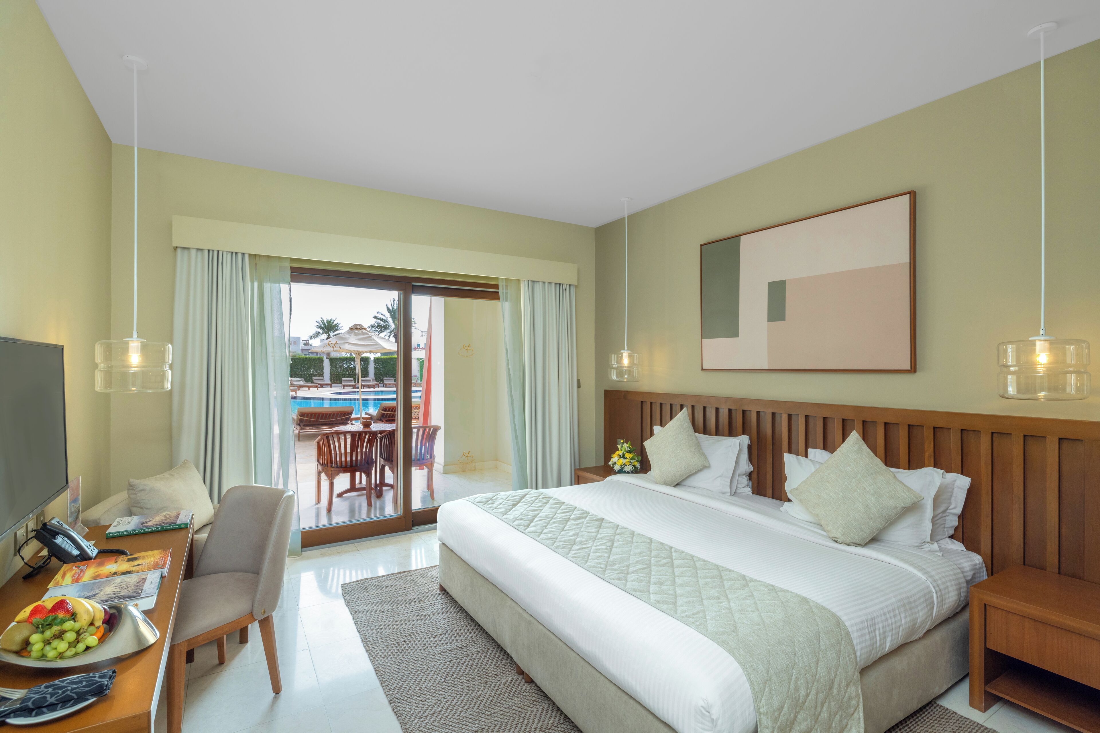 Une chambre d’hôtel avec un grand lit, un bureau avec un téléphone et une assiette de fruits, une télévision et une vue sur l’espace extérieur de la piscine.