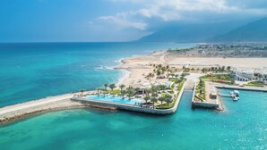 Vue aérienne d’une station balnéaire avec une piscine, une plage et des montagnes en arrière-plan.