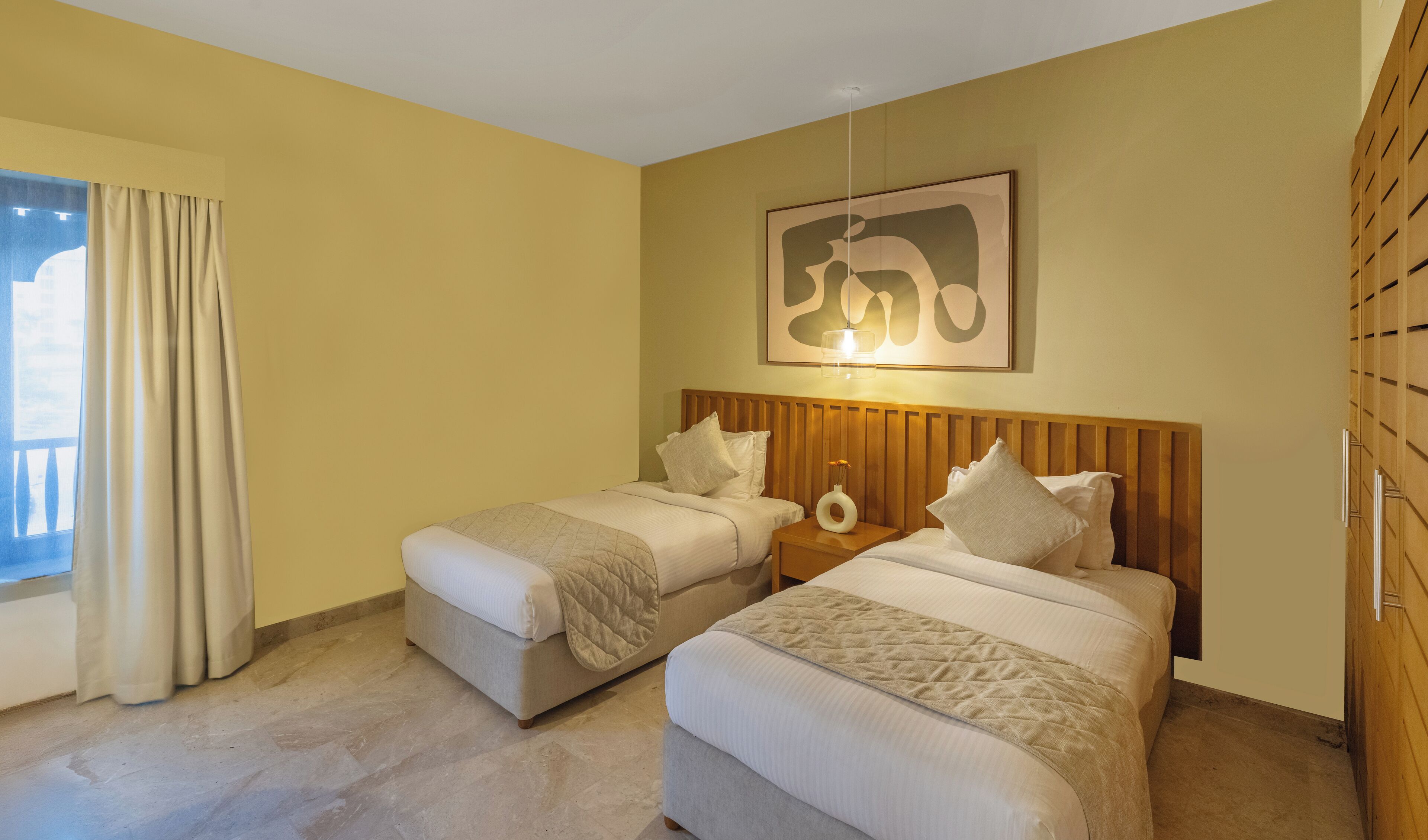 Une chambre d’hôtel avec deux lits, une tête de lit en bois, un tableau au mur et une fenêtre avec des rideaux.