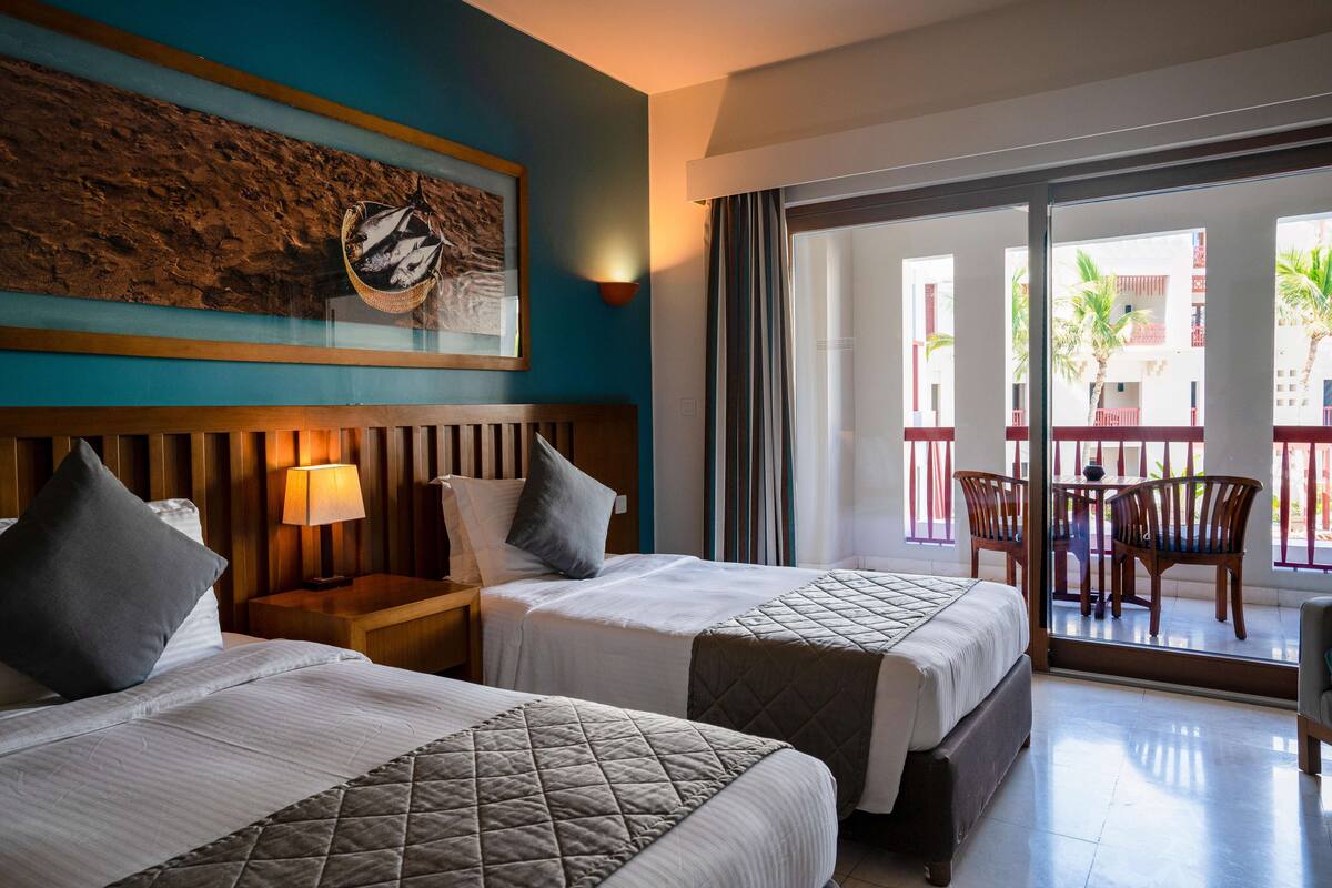 Une chambre d’hôtel avec deux lits, une tête de lit en bois, une œuvre d’art accrochée au mur, une table de chevet avec une lampe et un balcon avec une table et des chaises.