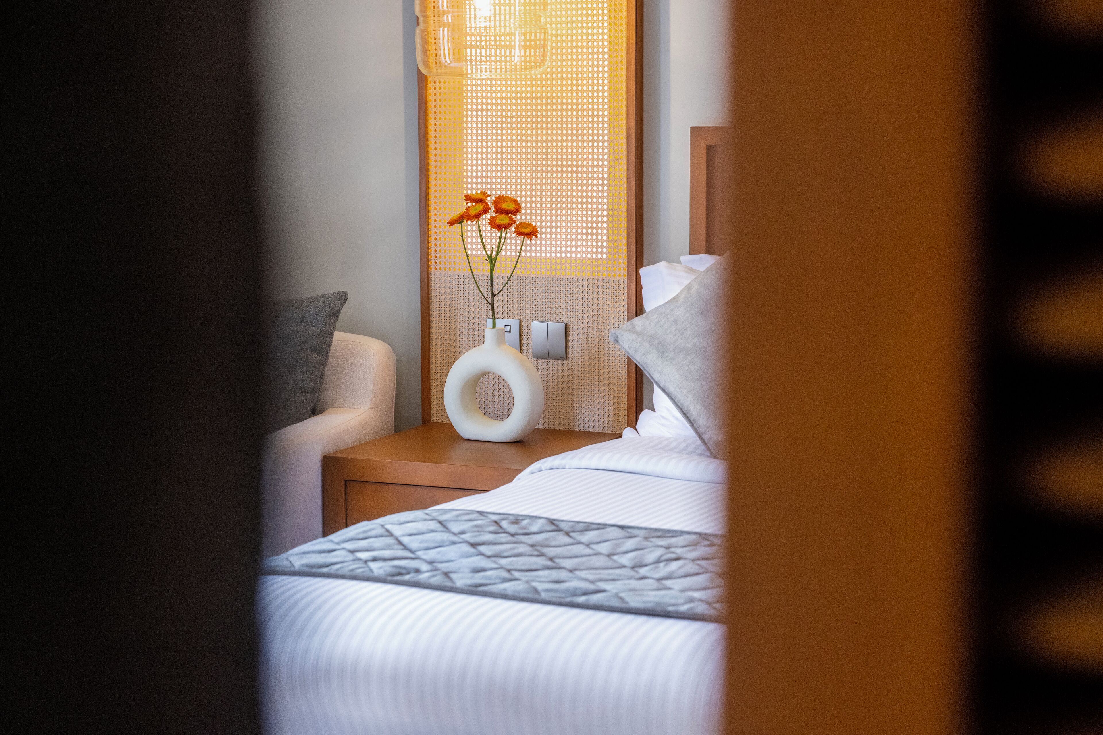 Un lit bien fait, recouvert d’une courtepointe matelassée, une table de chevet sur laquelle se trouve un vase blanc rempli de fleurs, et un luminaire fixé au mur.