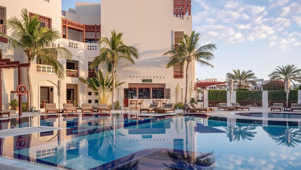 Une piscine entourée de chaises longues et de palmiers, le tout bordé d’un bâtiment doté de balcons et d’un espace bar.