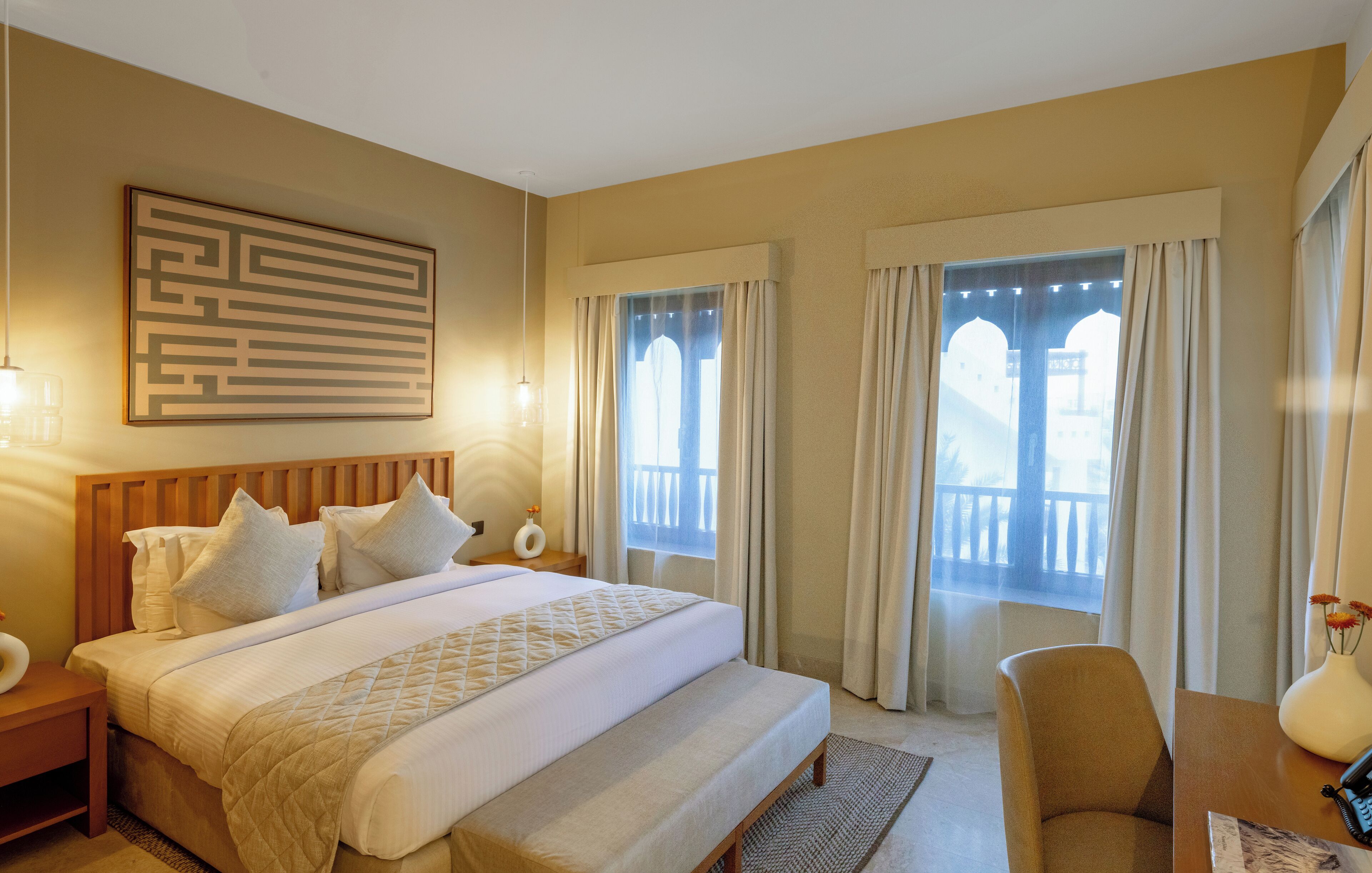 A hotel room with a large bed, a wooden headboard, a nightstand, a desk with a chair, a vase with flowers, and a window with curtains.