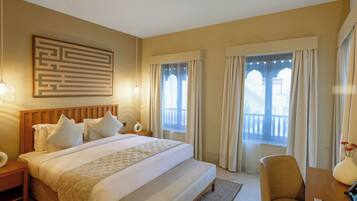 A hotel room with a large bed, a wooden headboard, a nightstand, a desk with a chair, a vase with flowers, and a window with curtains.