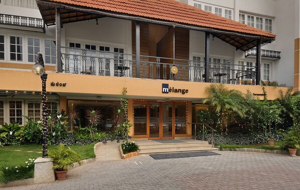 Ein Hotel mit auffälligem 'mélange'-Schild, einem überdachten Balkonbereich und einem gepflegten Garten mit Kübelpflanzen und einer Statue.