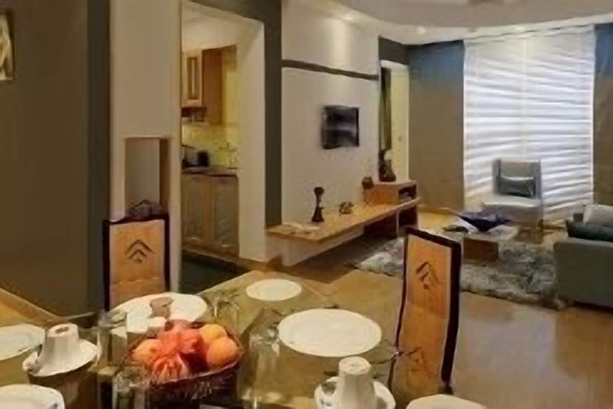 Un comedor con una mesa dispuesta para cuatro personas, una cesta de frutas y una cocina visible al fondo.
