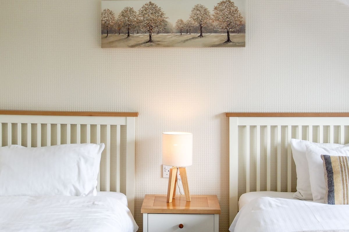 A hotel room with two beds, a nightstand with a lamp, and a painting of trees on the wall.