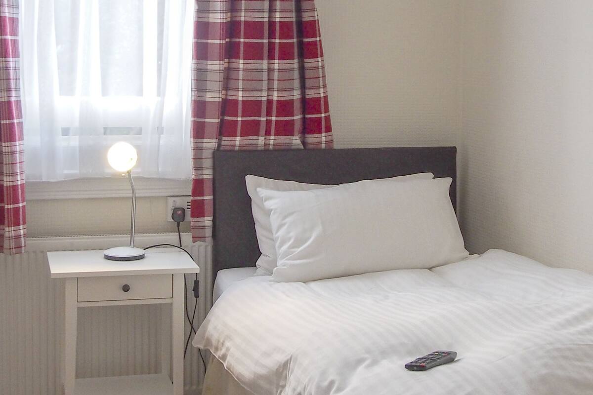 A bedroom with a bed, a bedside table with a lamp, a remote control on the bed, and a window with curtains.