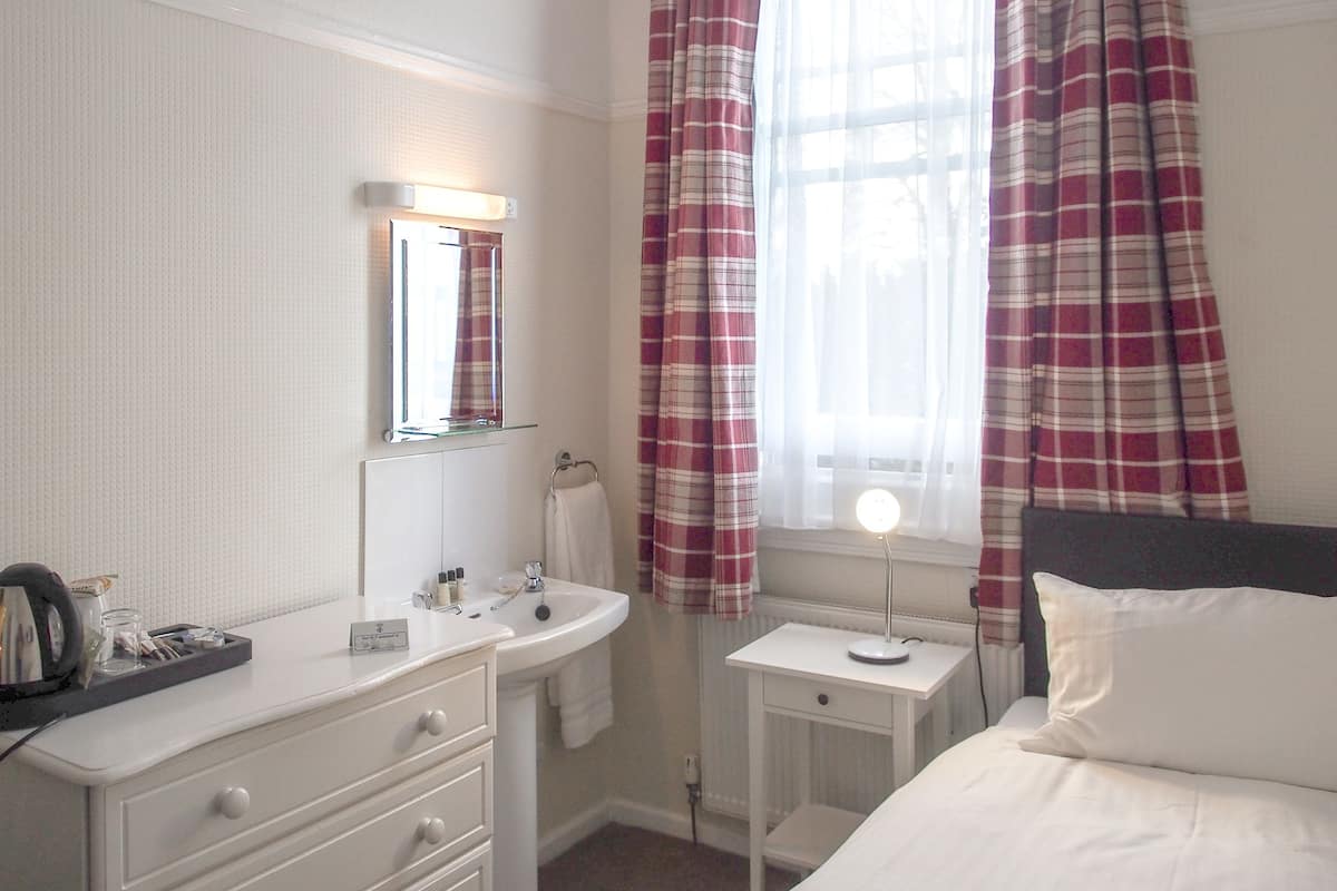 A hotel room with a white bed, a bedside table with a lamp, a white dresser with a mirror, a window with plaid curtains, and a radiator.