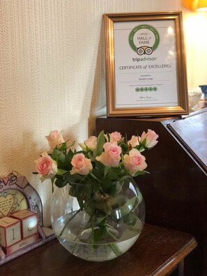 A framed TripAdvisor Hall of Fame certificate on a wall, a vase with pink roses on a wooden table, and a clock displaying the date 'April 10'. - Elmdon Lodge (Birmingham)