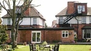 A two-story house with a brick facade, a visible chimney, and a small garden with a table and chairs. - Elmdon Lodge (Birmingham)