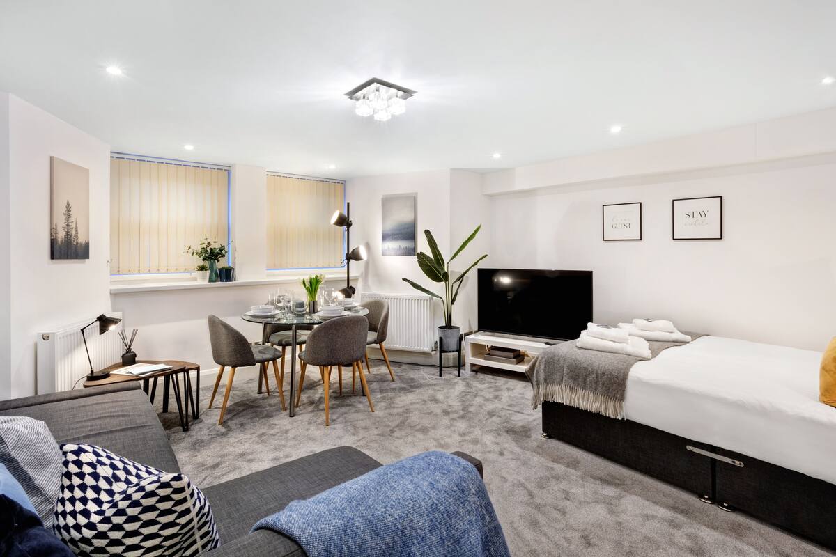 Ett modernt hotellrum med en soffa, ett matbord, stolar, en TV, en säng och ett stort fönster med gardiner.