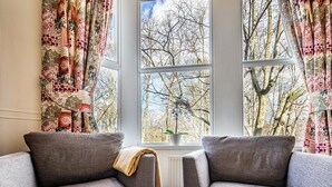 A cozy living room with a large window, a sofa, and a table with a book. - Fountains Guest House (Harrogate)