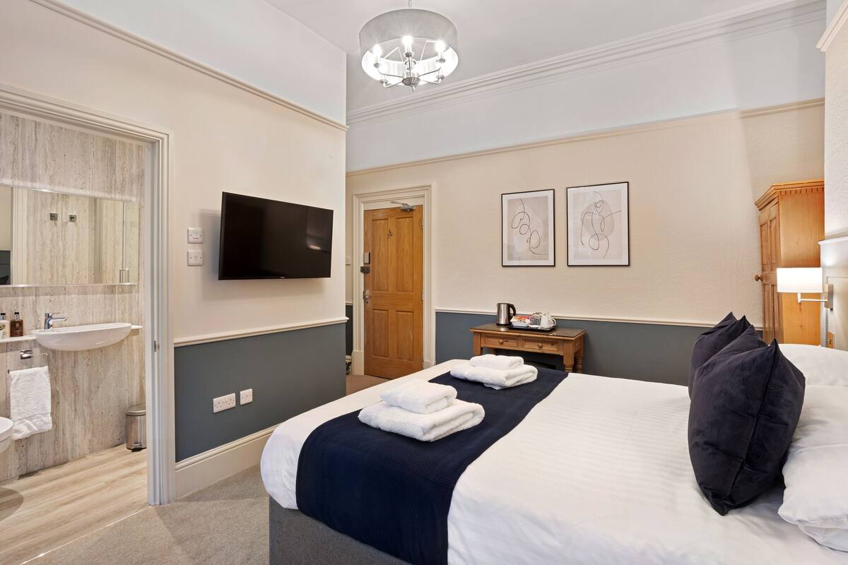 Ett hotellrum med en stor säng, en platt-TV, en trädörr och ett litet bord med en vattenkokare.