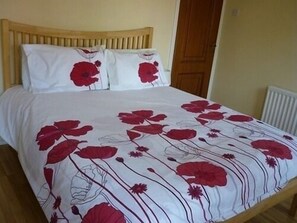 Una cama con colcha blanca de motivos florales rojos, cabecera de madera y dos almohadas blancas.