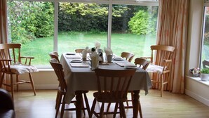 Un comedor con una mesa preparada para seis personas, rodeada de sillas de madera, ventanas amplias con cortinas y vistas a un jardín.