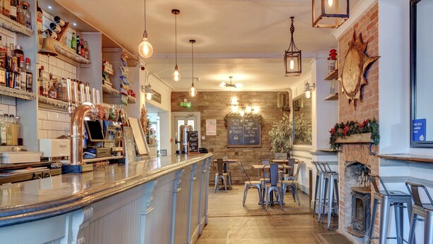 Uma área de bar com balcão, banquetas e prateleiras com garrafas. Uma parede de tijolos e um chão de madeira.