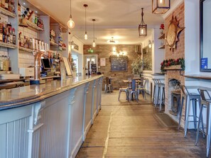 A bar area with a counter, stools, and shelves with bottles. There is a brick wall and a wooden floor.