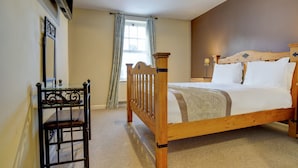 Quarto com cama de madeira, mesa de cabeceira pequena, uma cadeira, janela com cortinas e luminária no teto.