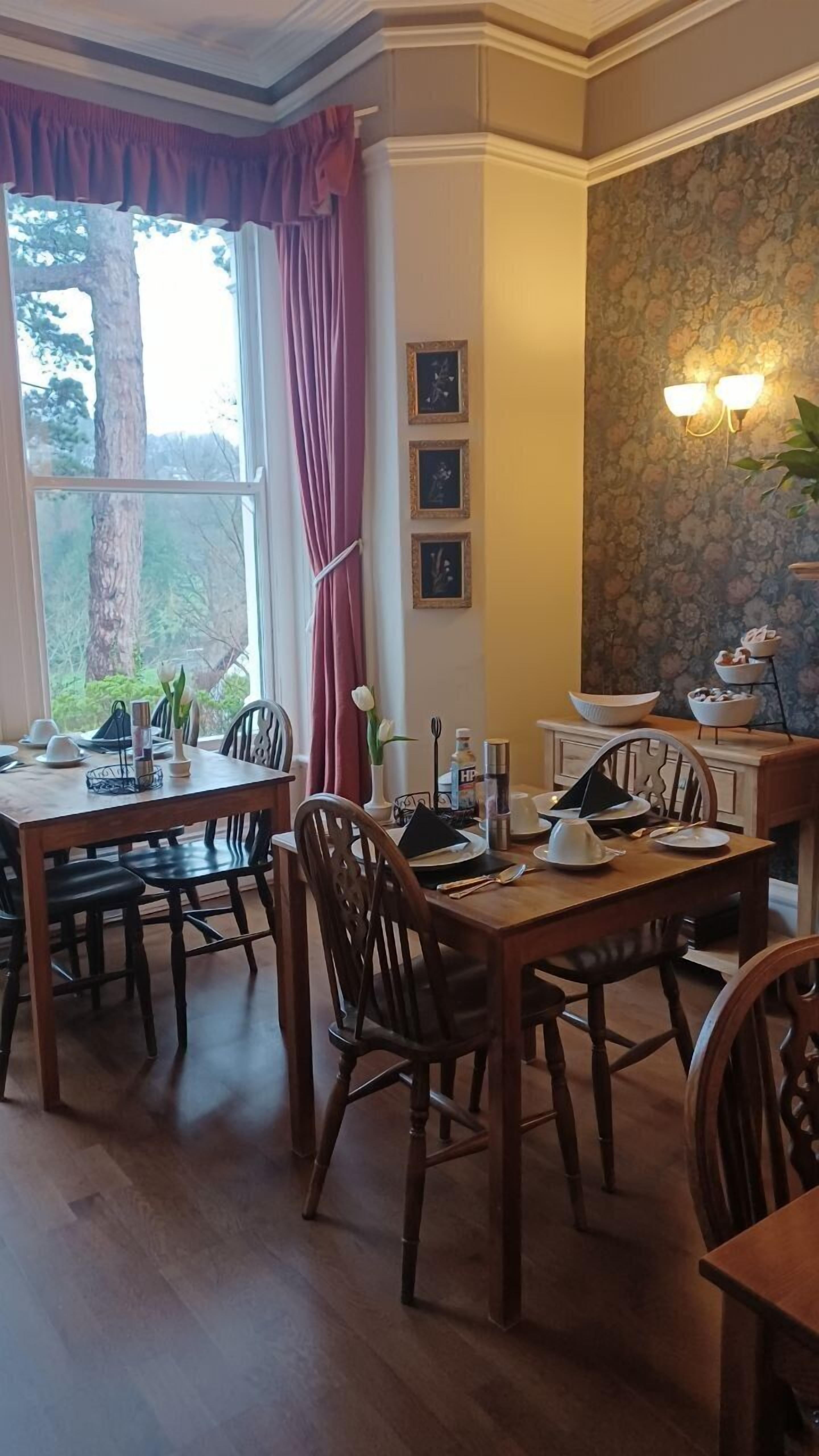 A dining area with a wooden table and chairs, a window with curtains, framed pictures on the wall, and a floral wallpaper pattern.
