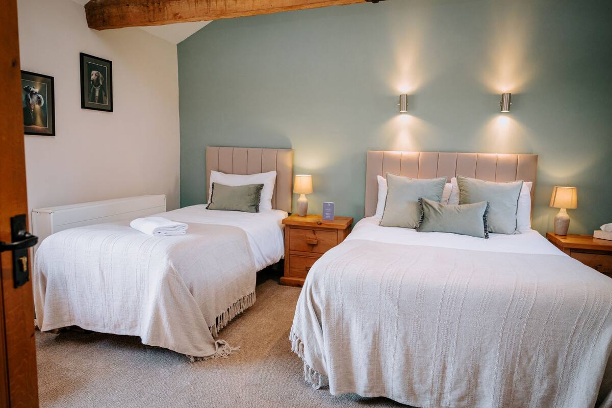 Ein Hotelzimmer mit zwei Betten, jeweils mit Kopfteil und Nachttischen. An der Wand befinden sich gerahmte Bilder und eine Holztür.