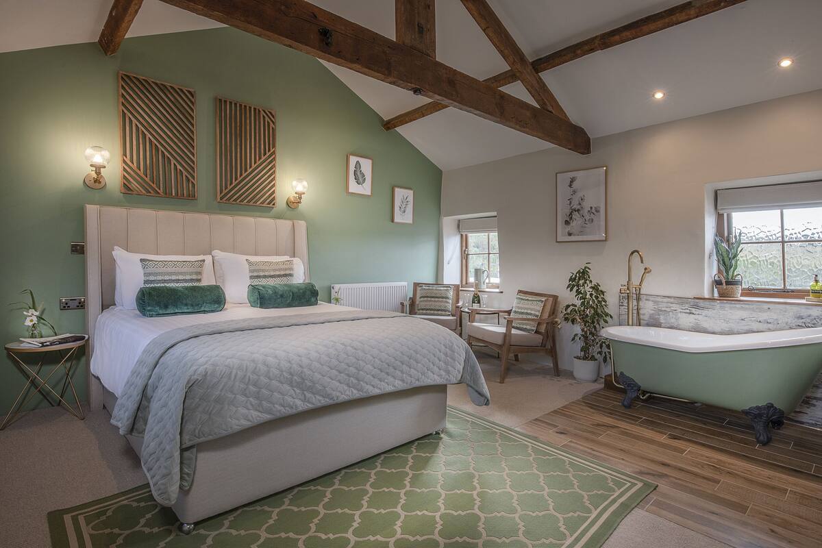 Ein Schlafzimmer mit einem großen Bett, einem grün gemusterten Teppich, Holzboden, einer grünen Badewanne und einem Fenster mit Aussicht.