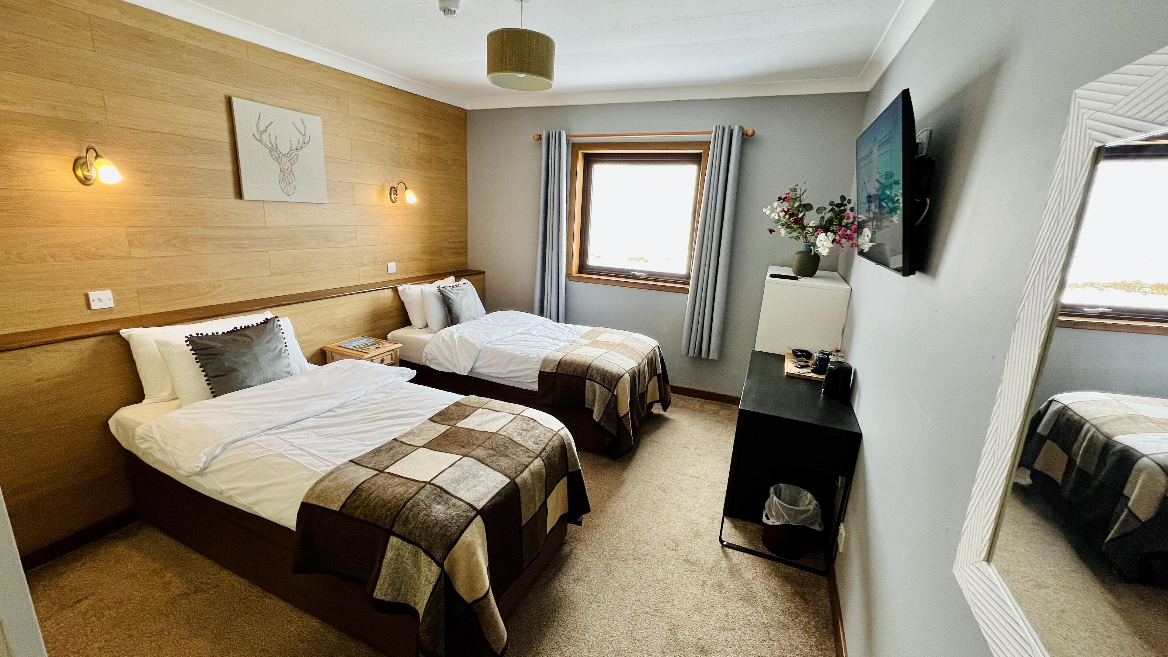 Une chambre d’hôtel avec deux lits, une tête de lit en bois, une fenêtre avec des rideaux, une petite table avec un vase et un téléviseur fixé au mur.