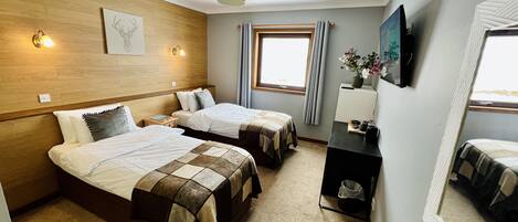 Une chambre d’hôtel avec deux lits, une tête de lit en bois, une fenêtre avec des rideaux, une petite table avec un vase et un téléviseur fixé au mur.