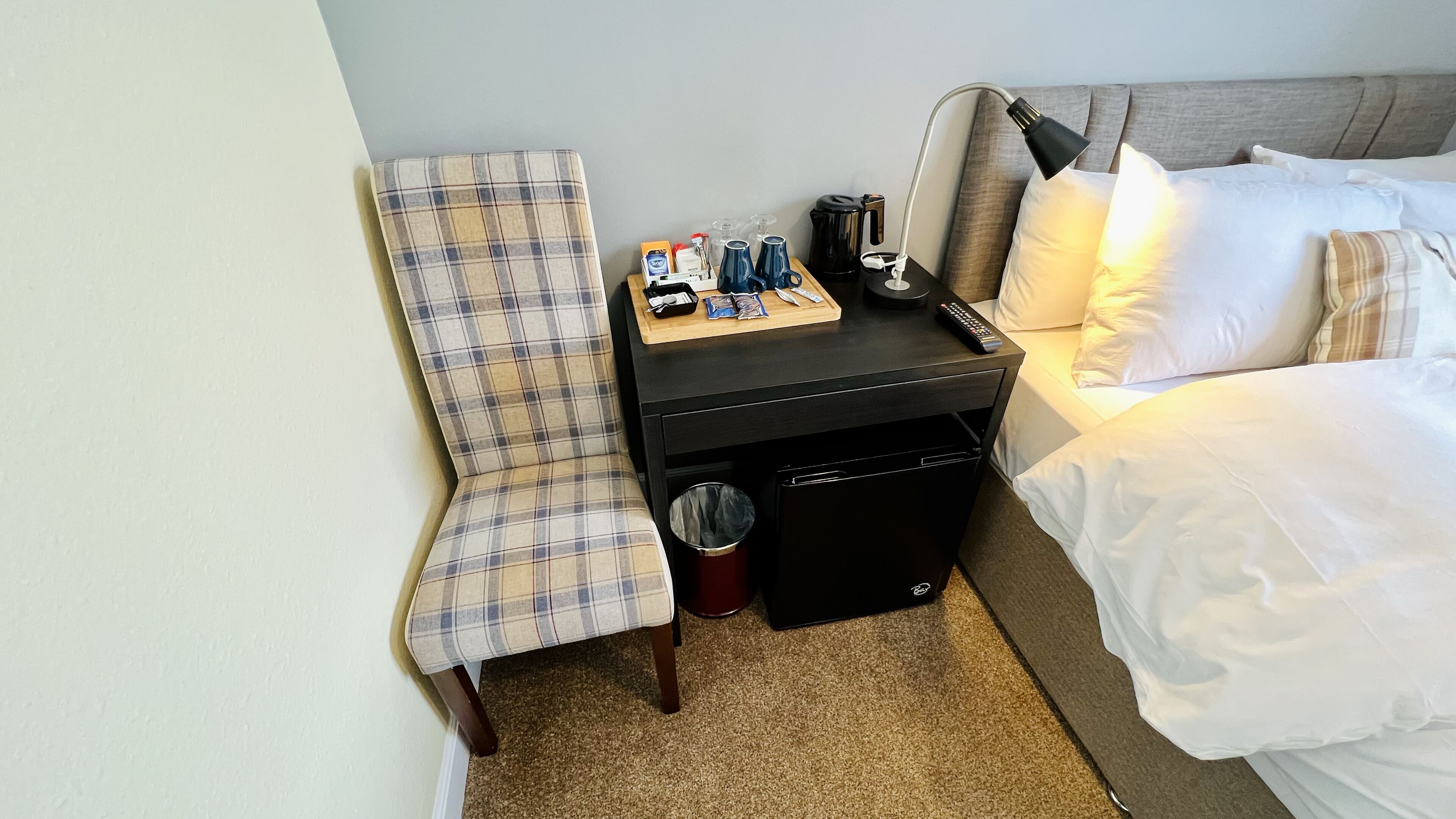 Una habitación de hotel con cama, una silla, una mesita con artículos de aseo, un cubo de basura y una mesita de noche con lámpara.