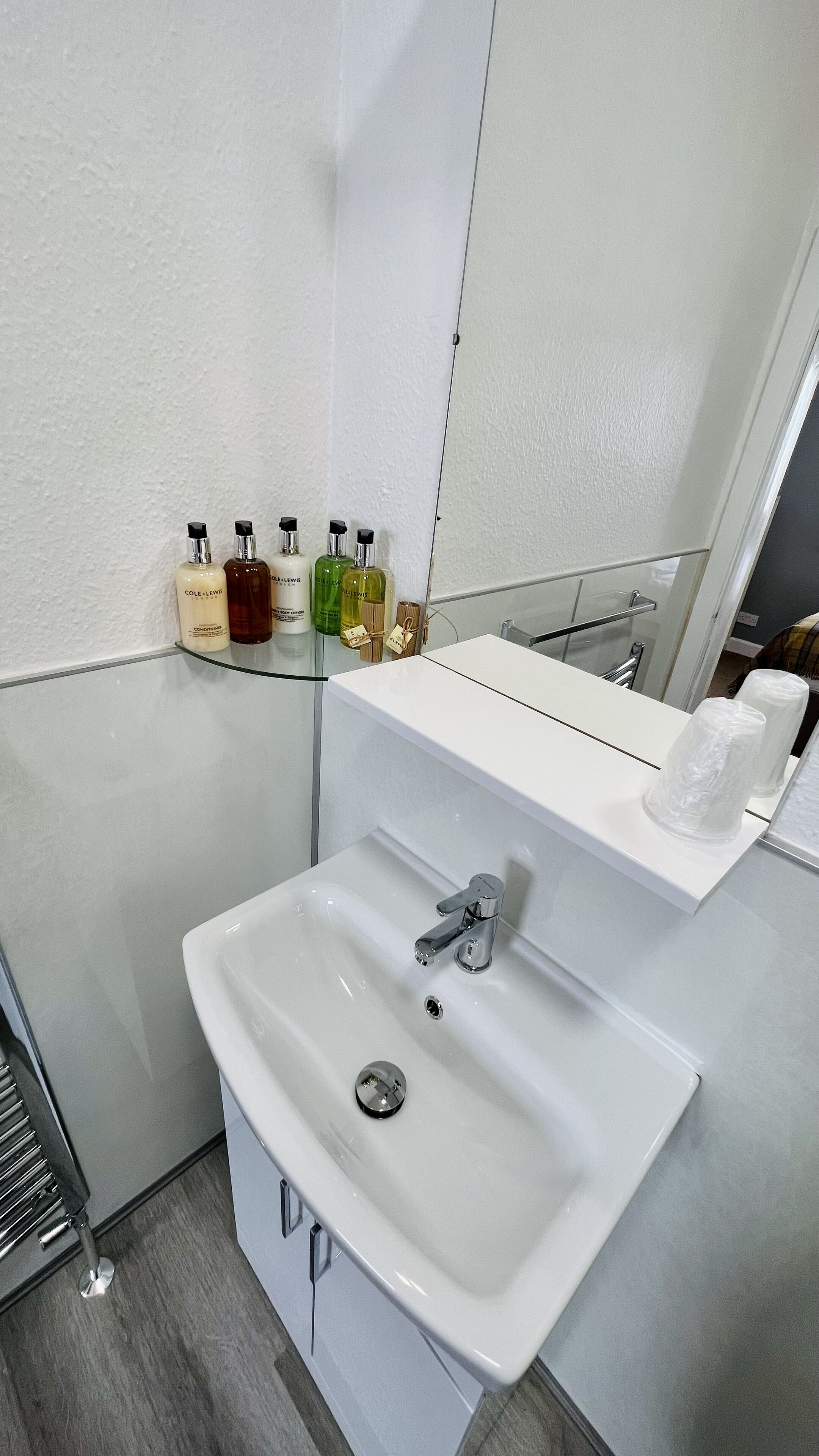 A bathroom with a white sink, chrome faucet, and a mirror above it. There are toiletries on a shelf and a towel rack.