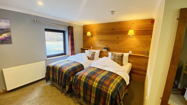 A hotel room with two beds, a window, a radiator, wooden paneling, and two wall-mounted lamps. - Aultguish Inn (Garve)