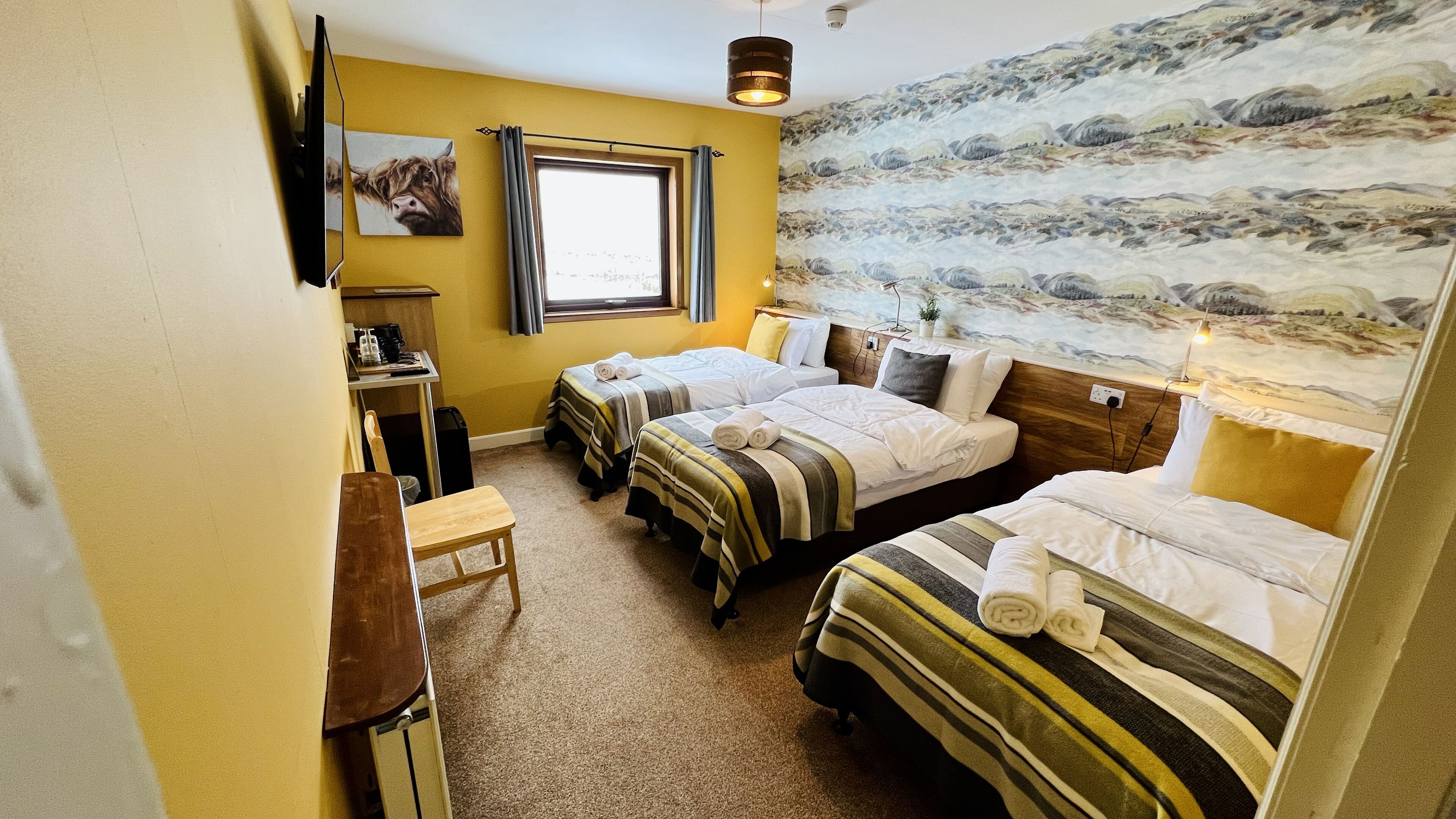 Habitación de hotel con dos camas individuales, un escritorio con silla, una mesita, un televisor montado en la pared y una ventana con cortinas.