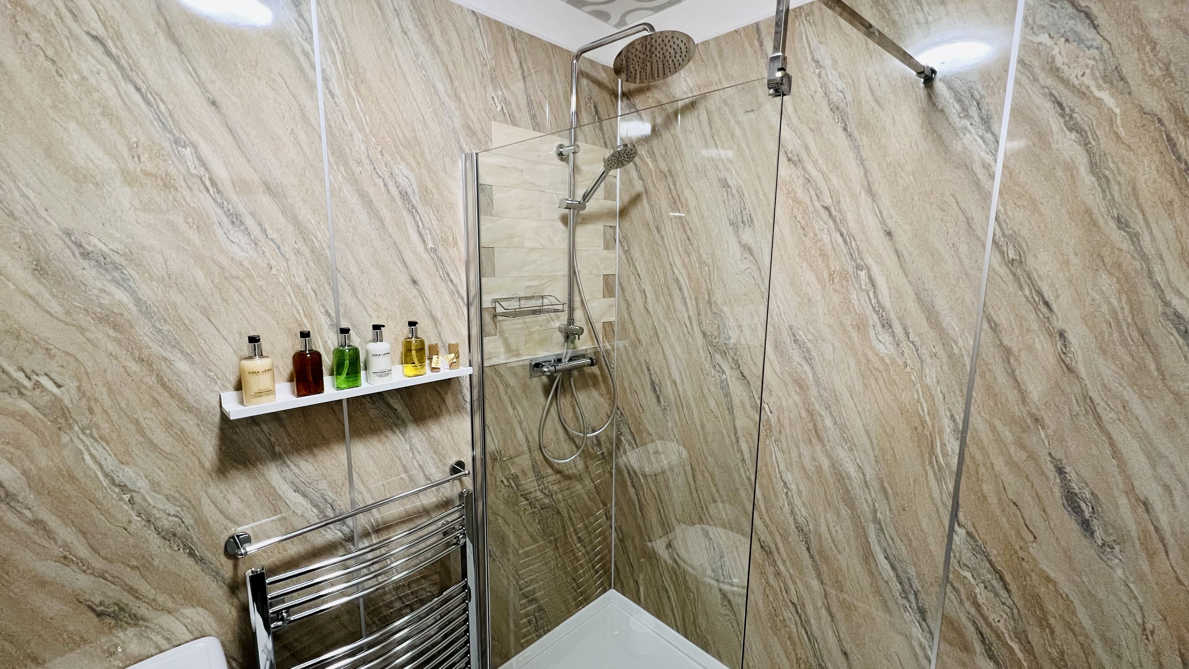 A modern bathroom with a glass-enclosed shower, marble walls, and a shelf with toiletries.